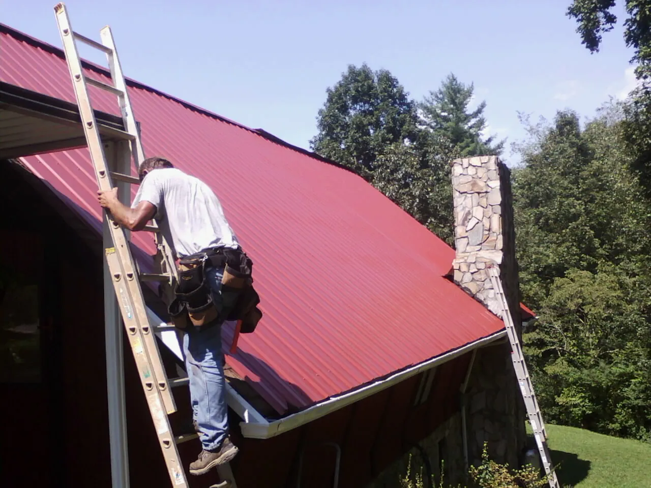 Roofer on a ladder beside a red metal roof and stone chimney — Soffit & Fascia Repair by Roofing Brothers in Moodus